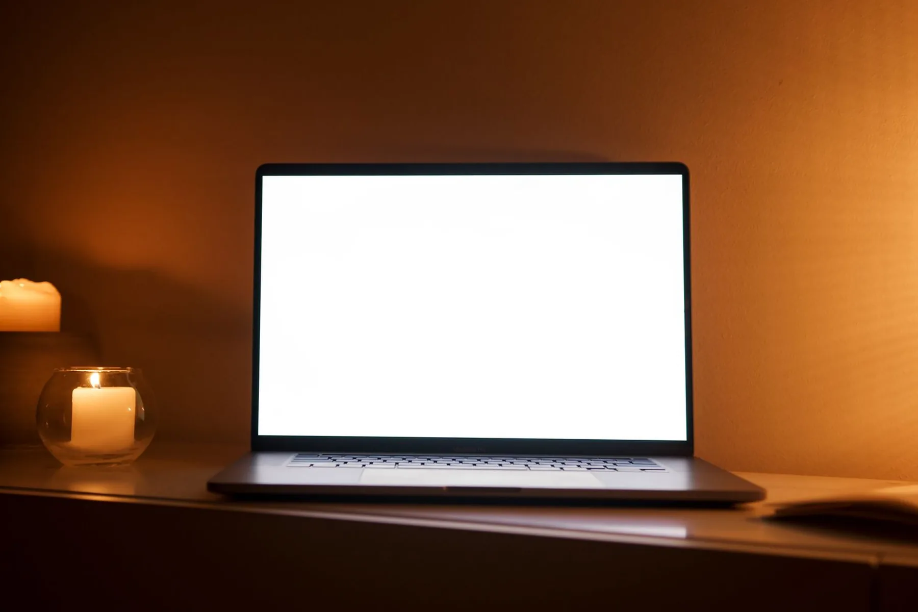 A laptop with a blank glowing screen beside a lit candle in a dark room