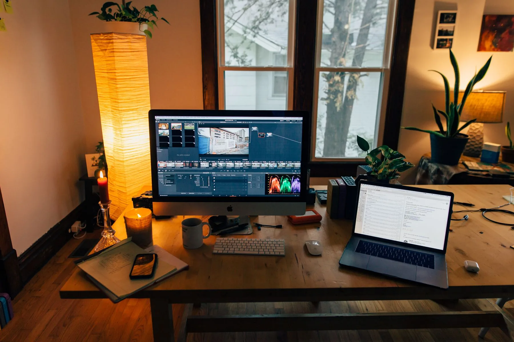 A warm home office at night with a desktop monitor, laptop, and a lit candle