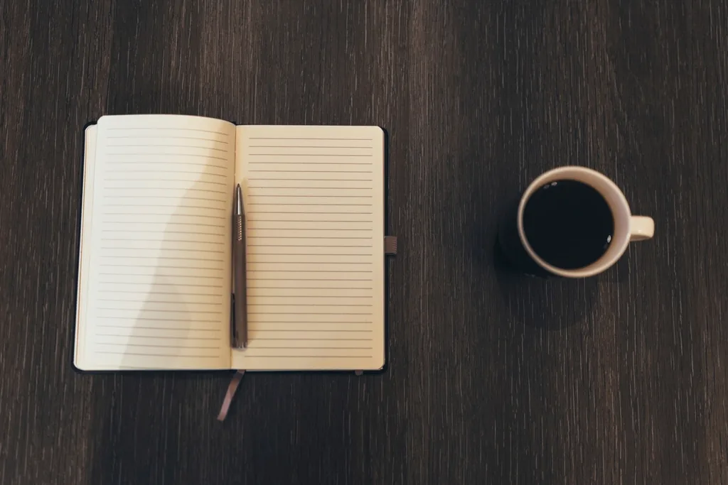 An open blank notebook with a pen beside a cup of black coffee on a dark wooden table