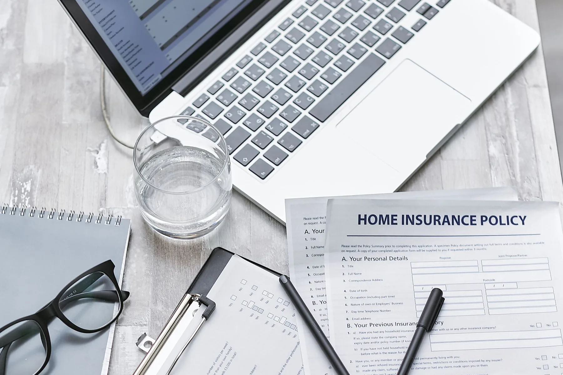 A home insurance policy, glasses, and a laptop on a light wooden desk